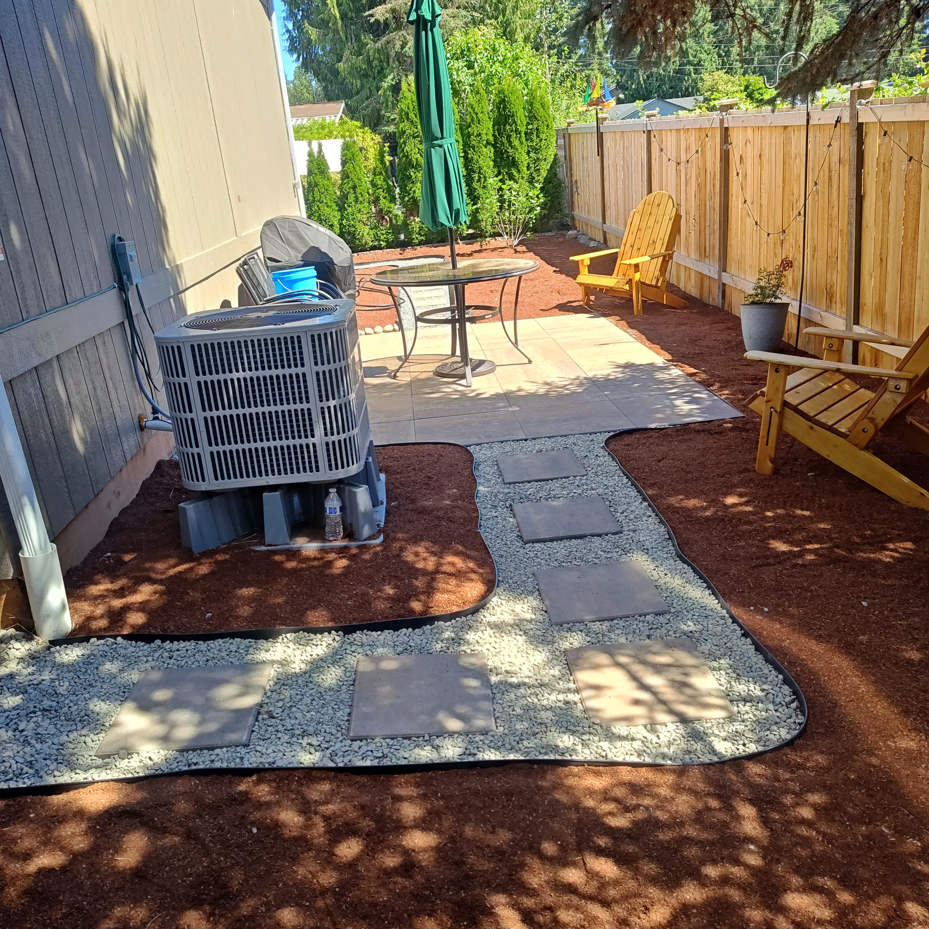 Backyard patio with stepping stones set in gravel pathways, red mulch beds, outdoor furniture, yellow Adirondack chairs, wooden fence with string lights.